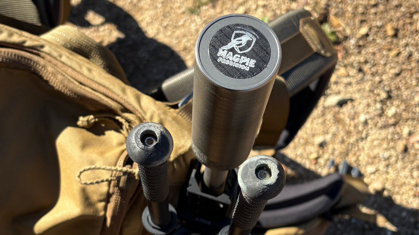 A close-up outdoor shot of a tactical bag with a mounted cylindrical metal accessory, likely a flashlight or camera light, featuring a visible 'Blackhawk' logo. A person's boot is visible at the bottom edge on a dirt ground surface.